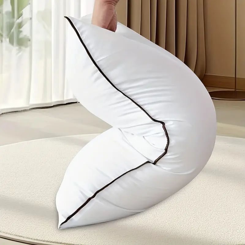 White U-shaped pillow with black piping held by a hand on a light-colored surface.